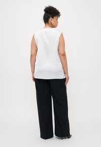 Top bianco senza maniche con una texture liscia, abbinato a pantaloni neri a gamba larga e scarpe con tacco, visto da dietro.