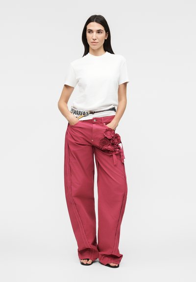 Γυναίκα που φοράει λευκό πουκάμισο με κοντά μανίκια, κόκκινο παντελόνι με φαρδιά μπατζάκια και floral εφαρμογή σε έναν μηρό, μαύρα σανδάλια και ζώνη με σχέδιο.