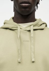 Κοντινό πλάνο ατόμου που φοράει ανοιχτό μπεζ φούτερ με χοντρές κορδόνια και ανάγλυφο λογότυπο "HUGO" στο κέντρο του στήθους.