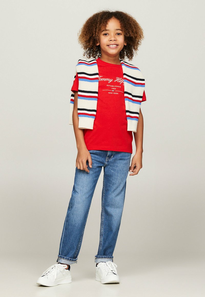 T-shirt rossa in cotone con logo "Tommy Hilfiger", abbinata a jeans blu chiaro e un gilet lavorato a maglia a righe crema drappeggiato sulle spalle. Sneakers bianche.