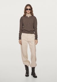 Brauner Strickpullover mit V-Ausschnitt und hellfarbigem Kragen, kombiniert mit beigen weit geschnittenen Hosen und schwarzen Stiefeletten.