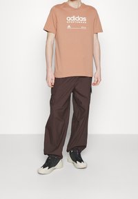 Světle hnědé bavlněné tričko s bílým logem "Adidas Sportswear", v kombinaci s tmavě hnědými cargo kalhotami a černými sportovními botami se šedými akcenty.