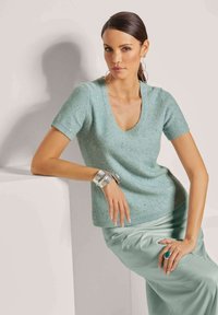 Lichtgroene korte mouw trui met een V-hals, voorzien van een textuur en glinsterende afwerking. Gecombineerd met een satijnen mintgroene rok en zilveren sieraden.