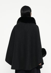 Svart ullkappe med pelskrage, med rund nederkant og teksturerte detaljer. Bakdelen har et elegant, minimalistisk design.