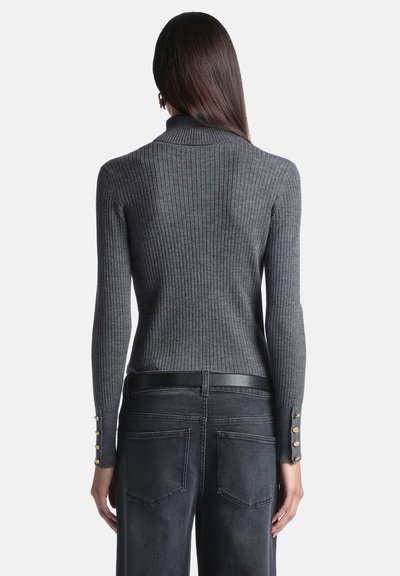 Femme aux longs cheveux foncés porte un pull gris côtelé ajusté et un jean noir taille haute, vue de dos sur fond blanc.