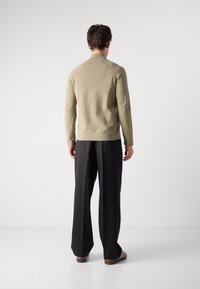 Man med ljusbeige texturerad långärmad skjorta, svarta vida byxor och bruna skor som står vänd mot en helt vit vägg.