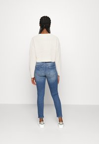 Maglione all'uncinetto beige, design corto; jeans in denim blu con una leggera sfumatura; sneakers bianche con accenti grigi e logo sul tallone.