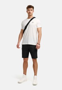 Hvid T-shirt med korte ærmer, sorte shorts og hvide høj-top sneakers. Inkluderer en sort crossbody taske. Modellen står oprejst mod en neutral baggrund.