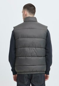 Grau gepolsterte Weste mit hohem Kragen und horizontalen Nähdetails. Getragen über einem dunklen Pullover, der ein sauberes, minimalistisches Design zeigt.