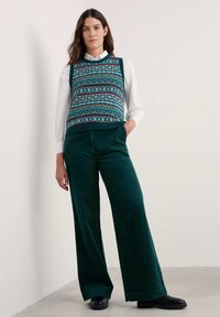 Chaleco de punto con patrones geométricos en azul verdoso, blanco y burdeos, superpuesto a una camisa blanca; combinado con pantalones de pana de pierna ancha en verde oscuro.