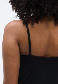 Μαύρο τοπ με λεπτές, ρυθμιζόμενες τιράντες, λεία υφή και διακριτική γυαλάδα, που αναδεικνύει στρογγυλεμένη πλάτη και μινιμαλιστικό σχέδιο.