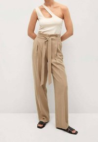 Osoba nosząca beżowe spodnie z wysokim stanem i szerokimi nogawkami, z paskiem zawiązanym z przodu, białą bluzkę na jedno ramię i czarne sandały na obcasie z odkrytymi palcami.