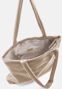 Sac cabas en cuir matelassé beige avec deux longues poignées, fermeture éclair et une poche intérieure zippée, doté d'une doublure en tissu lisse.