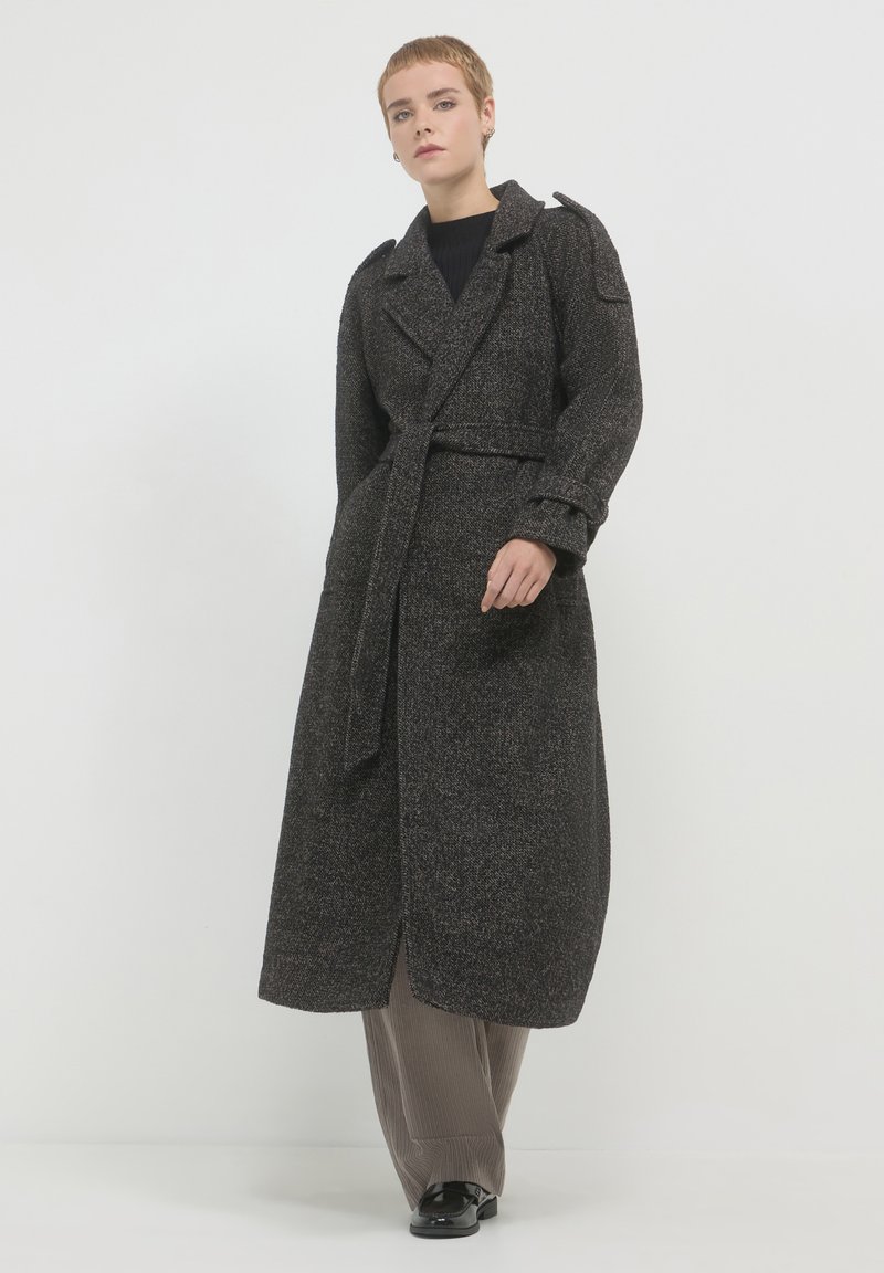 Lungo cappotto nero texturizzato con vita cinturata, ampi rever e spalline, abbinato a pantaloni marroni plissettati e scarpe nere.