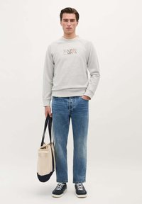Grå sweatshirt med "SCALPERS COMPANY" trykt i farver, parret med blå jeans og sorte sneakers. Holder en beige og marineblå taske.