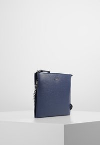 Granatowa torebka DKNY z teksturowanej skóry, o kwadratowym kształcie, zamykana na suwak u góry, z akcentem w postaci łańcuszka. Minimalistyczny design bez dodatkowych ozdób.