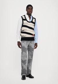 Un gilet tricoté noir et crème rayé superposé à une chemise bleu clair, associé à un jean gris arborant un léger motif angélique. Chaussures noires.
