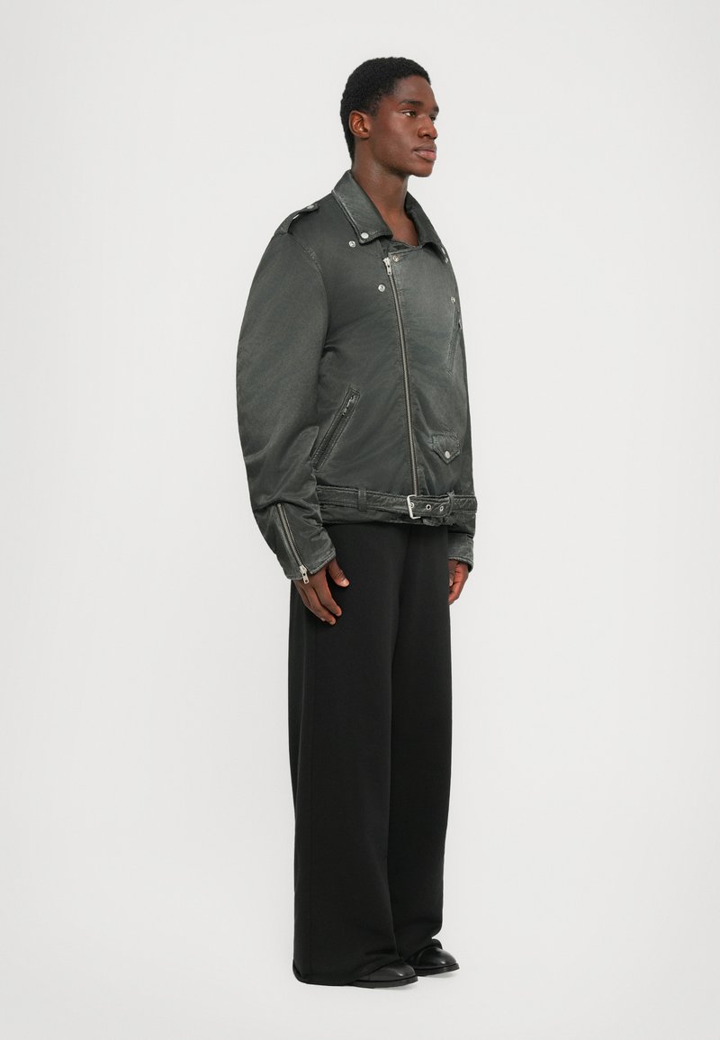 Black oversized denim jacket with a zip front, shoulder epaulettes, and two side pockets, worn with wide black trousers.
