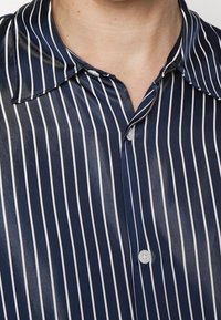 Cou et haut de la poitrine portant une chemise en satin bleu marine à fines rayures blanches verticales et boutons blancs, col ouvert en haut.