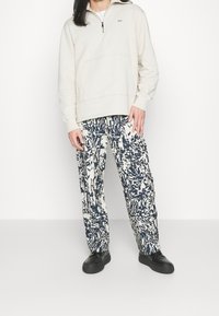 Béžová mikina na zip s přední kapsou, doplněná o tmavě modré a bílé kalhoty s abstraktním vzorem. Černé nazouvací boty dokončují outfit.