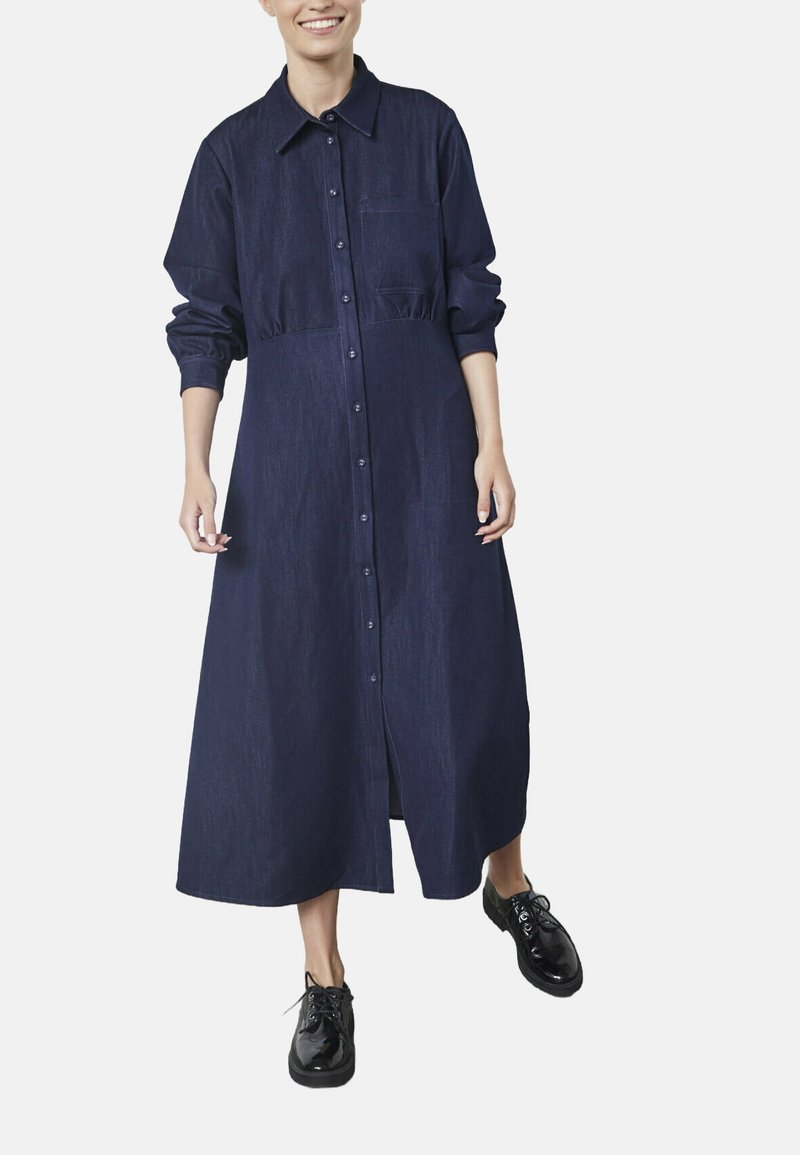 Denim skjortekjole i mørkeblå, med knaplukning foran, lange ærmer med elastiske manchetter, krave og en frontlomme. Sammen med sorte sko.