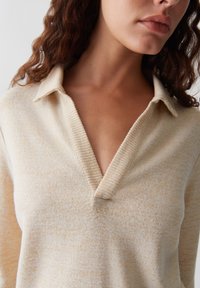 Woman with wavy brown hair wearing a beige V-neck sweater with a collar, showing her neck and lower face against a plain background.