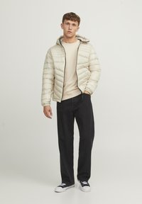 Light beige puffer jacket with horizontal quilted design, front zipper, and adjustable hood. Worn over a cream sweater and black pants.