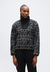 Γυναίκα με αλογοουρά που φοράει μαύρο και άσπρο φλις τζάκετ Columbia με σχέδιο και μαύρο παντελόνι, στέκεται μπροστά σε απλό ανοιχτό φόντο.