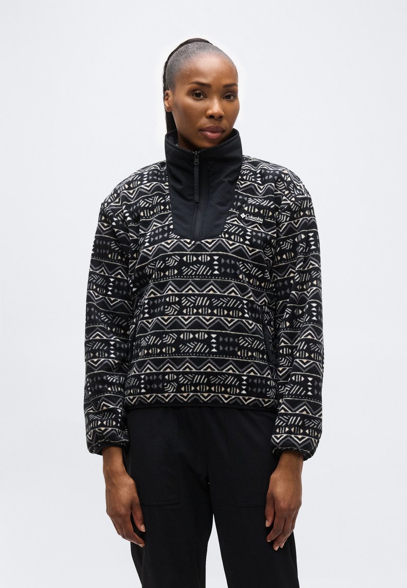 Γυναίκα με αλογοουρά που φοράει μαύρο και άσπρο φλις τζάκετ Columbia με σχέδιο και μαύρο παντελόνι, στέκεται μπροστά σε απλό ανοιχτό φόντο.