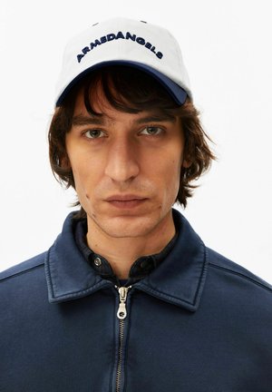 Junger Mann mit mittellangen braunen Haaren, der eine weiße und marineblaue Mütze mit dem Schriftzug "ARMEDANGELS" und eine marineblaue Jacke mit Reißverschluss vor einem weißen Hintergrund trägt.