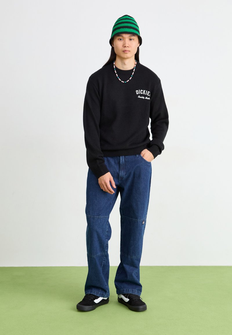 Pull noir avec le logo "Dickies", jean bleu, baskets noires, bonnet rayé vert, et un collier de perles colorées. Debout sur un sol vert.