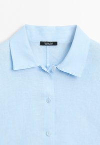 Col de chemise bleu clair à boutons et partie avant supérieure avec trois boutons visibles et étiquette noire portant la mention Massimo Dutti fabriqué au Maroc.