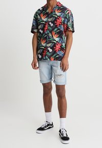 Camisa hawaiana negra de manga corta con estampado floral tropical colorido; combinada con pantalones cortos de denim rasgados de color azul claro y zapatillas deportivas negras.