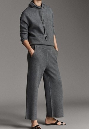 Personne debout, les mains dans les poches, portant un sweat à capuche gris foncé, un pantalon large assorti et des sandales noires à bouts ouverts.