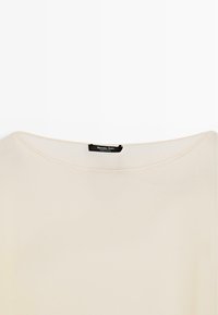 Kremfarvet skjorte med en bred rund halsudskæring og et sort mærke på bagsiden af kraven med påskriften "Massimo Dutti Made in China".