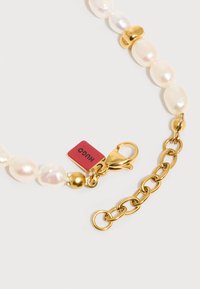 Κολιέ από πέρλες με χρυσούς κρίκους αλυσίδας, ένα χρυσό κούμπωμα σε σχήμα αστακού και μια μικρή κόκκινη τετράγωνη ετικέτα με χαραγμένο το "HUGO."