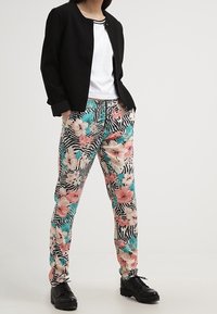 Veste noire légère sur une chemise blanche, associée à un pantalon à motifs avec un design floral rose et bleu-vert sur fond rayé. Chaussures noires.