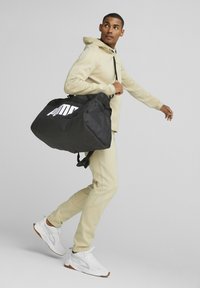 Bolsa de deporte negra con un gran logotipo blanco, cuenta con una parte superior con cremallera y una correa de hombro ajustable, llevada por una persona con un atuendo beige.