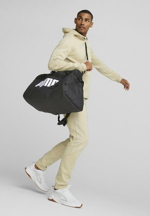 Bolsa de deporte negra con un gran logotipo blanco, cuenta con una parte superior con cremallera y una correa de hombro ajustable, llevada por una persona con un atuendo beige.