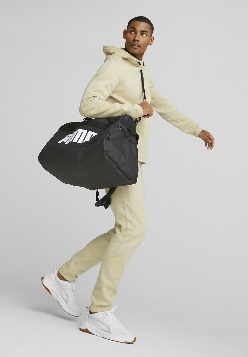 Bolsa de deporte negra con un gran logotipo blanco, cuenta con una parte superior con cremallera y una correa de hombro ajustable, llevada por una persona con un atuendo beige.