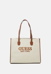 Canvas tote bag in cream with brown faux leather accents, featuring "GUESS EST 1981" logo in orange. Dual handles for easy carrying.