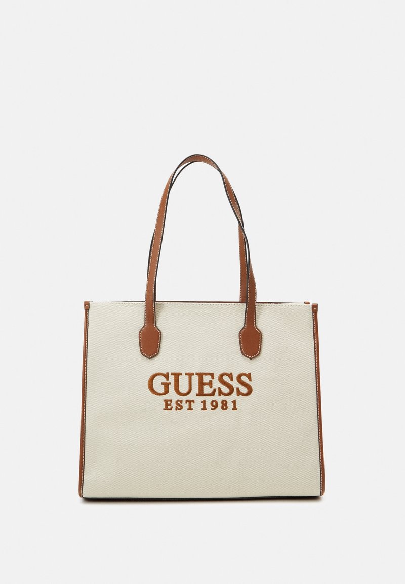 Canvas tote bag in cream with brown faux leather accents, featuring "GUESS EST 1981" logo in orange. Dual handles for easy carrying.
