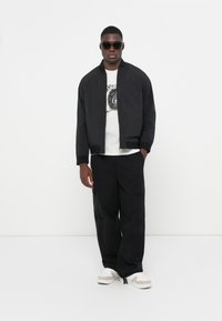 Black bomber jacket, white graphic t-shirt, black cargo pants, and white shoes with beige accents. Sunglasses complete the look.