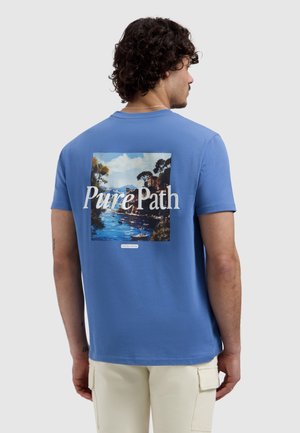 Man met de rug naar de kijker, draagt een blauw T-shirt met een landschapsschilderij en de tekst "Pure Path" boven water en bomen, gecombineerd met lichte cargobroek.