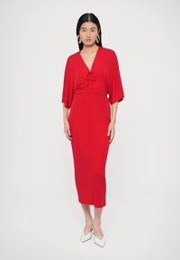 Červené midi šaty s netopierími rukávmi, záhybmi v páse a hladkou textúrou. Doplnkové biele lodičky so špicatou špičkou.
