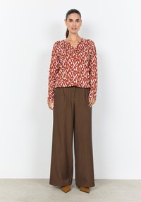 Rot-weiß gemusterte Bluse mit langen Ärmeln, kombiniert mit braunen, weit geschnittenen Hosen; das Modell trägt beigefarbene, spitz zulaufende Schuhe; glatte Texturen sind deutlich erkennbar.