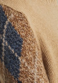 Pull en maille avec une base beige et un panneau texturé de motifs géométriques bleus et marron sur l'épaule, mettant en valeur des fils variés.
