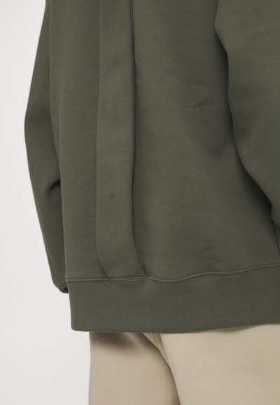 Hanorac de culoare verde măsliniu, cu o textură moale, având un detaliu de cusătură laterală și manșete ribbate la margine, asortat cu pantaloni bej.