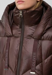 Braune gesteppte Steppjacke mit hohem Kragen, ausgestattet mit einem Frontreißverschluss und Logo-Bändern. Glatte glänzende Textur mit dezenten Marken-Details.