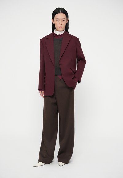 Blazer oversize color marrone scuro con spalle strutturate, indossato sopra un maglione scuro, abbinato a pantaloni larghi marroni e scarpe bianche a punta.